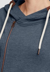 Sudadera con capucha en mezcla de algodón azul oscuro, con cremallera frontal, cordones beige con acentos marrones y una textura suave.