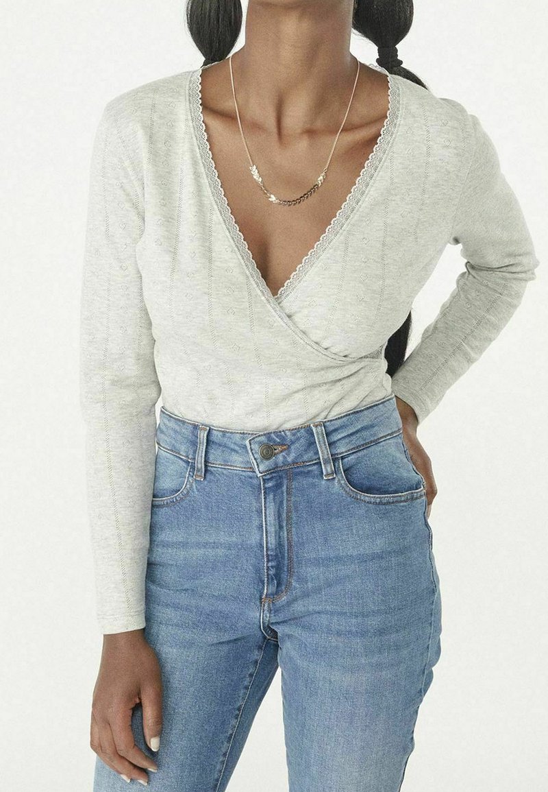 Hellgraues langärmliges Wickeloberteil mit tiefem V-Ausschnitt und Spitzenbesatz, kombiniert mit mittelhohen blauen Jeans aus Denim im klassischen Fünf-Taschen-Design.