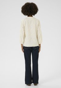 Balta siuvinėta blusa su pūstomis rankovėmis, tekstūrinis gėlių raštas, derinama su tamsiai mėlynomis platėjančiomis džinsinėmis kelnėmis. Iš nugaros matosi audinio detalė.