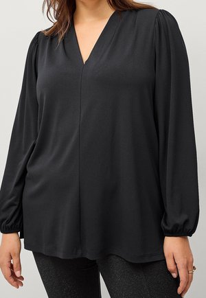 Kobieta w czarnej bluzce z długim rękawem z marszczonymi ramionami i elastycznymi mankietami, w połączeniu z błyszczącymi czarnymi spodniami.