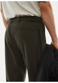Dark green trousers with a smooth texture, elastic waistband, and a back pocket. A black jacket is held in the right hand.
