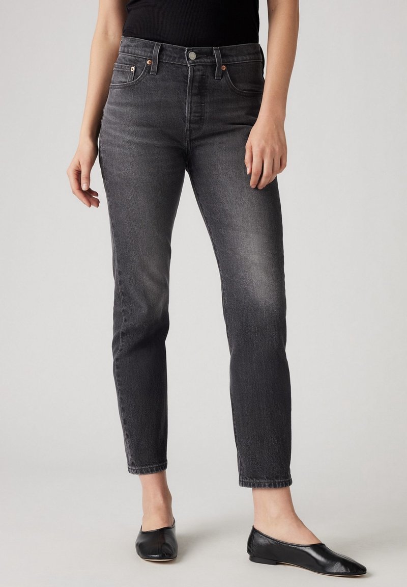 Femme portant un jean slim noir délavé associé à des ballerines noires, debout devant un fond gris clair uni.