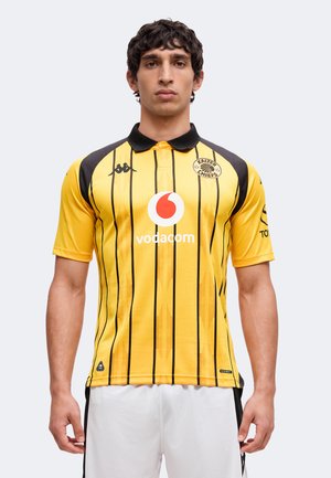 Homme portant un maillot de football rayé jaune et noir des Kaizer Chiefs avec le logo Vodacom, debout devant un fond clair uni.