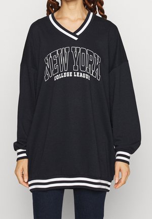 Osoba nosząca czarną, oversize'ową koszulę z długim rękawem z białymi paskami na kołnierzu, mankietach i na dole, z napisem „NEW YORK COLLEGE LEAGUE” z przodu.