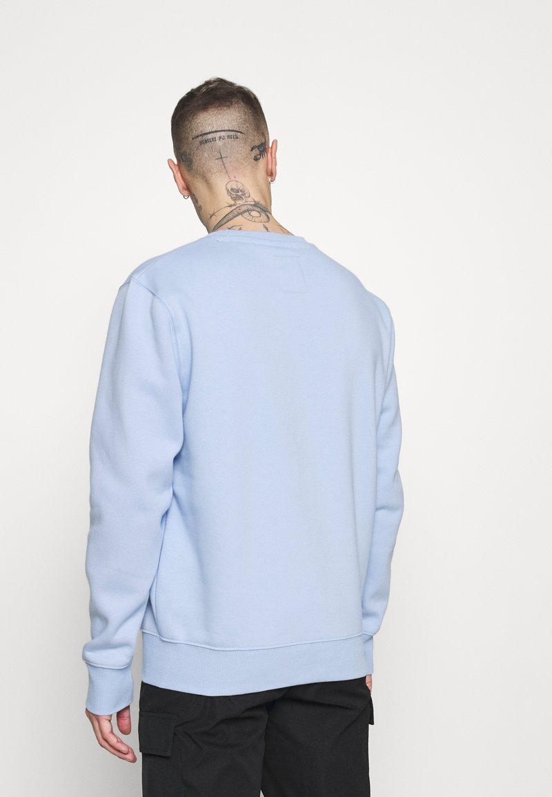 Felpa azzurra chiara con collo a giro, polsini e fondo a costine, texture morbida e un piccolo tag sul retro, indossata su pantaloni cargo scuri.