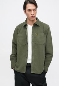 Camisa verde oliva de botones con tejido texturizado, dos bolsillos en el pecho y un pequeño logo amarillo en el bolsillo izquierdo. Llevada sobre una camiseta blanca.