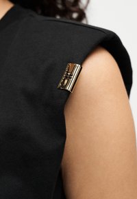 Μαύρο κορμάκι από βαμβάκι με δομημένους ώμους, διακοσμημένο με γυαλιστερό χρυσό κούμπωμα στο μανίκι. Ομαλή υ φή, μινιμαλιστικός σχεδιασμός.