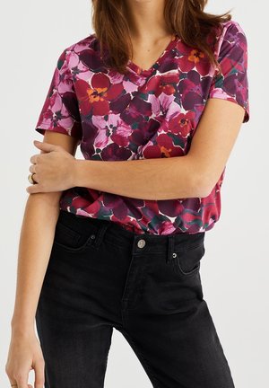 T-shirt à col en V à motifs floraux dans des tons de rose et violet, présentant un motif de grandes fleurs sur un tissu léger. Associé à un jean noir.