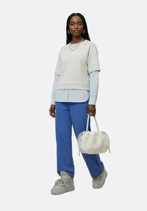 Mujer con largas trenzas lleva un jersey gris claro, pantalones azules, zapatos grises y sostiene un bolso blanco peludo, de pie frente a un fondo liso.