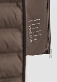 Section de veste marron avec fermeture éclair affichant l'étiquette Pako Lorente indiquant les caractéristiques : déperlante, coupe-vent, durable, isolation thermique et compressible.