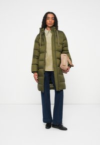 Abrigo acolchado verde oliva con capucha, que presenta un cierre frontal y cordones ajustables. Lleva puesto sobre una camisa beige clara y unos vaqueros de pierna ancha azul oscuro.