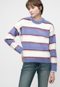En ung person iført en ullen stripete genser i blått, hvitt og rødt, kombinert med blå jeans, og poserer med en hånd på hoften mot en hvit bakgrunn.