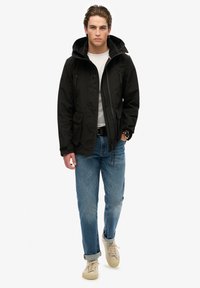 Giacca nera impermeabile con cappuccio, chiusura con zip, due tasche frontali e dettagli del logo in rilievo, abbinata a jeans azzurri chiari e sneakers beige.