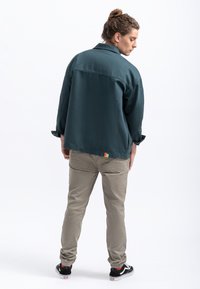 Giacca camicia in teal scuro con colletto, vestibilità rilassata e un piccolo etichetta multicolore all'orlo. I pantaloni beige e le sneakers nere completano il look.
