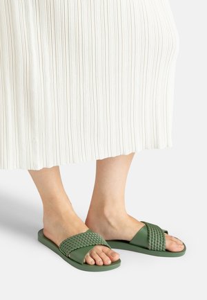Füße tragen grüne Slide-Sandalen mit geflochtenen Riemen, gezeigt mit einem weißen, gerippten Rock, der ab Mitte Wade sichtbar ist, vor einfachem Hintergrund.