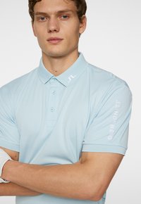 Ljusblå pikétröja med krage, korta ärmar och en subtil logotyp på kragen. Har en knappslå och en figursydd design.