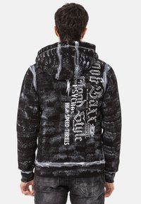 Schwarzer Hoodie mit einem strukturierten Muster und weißen Pinselstrichen. Auf der Vorderseite ist kräftiger Grafiktext abgebildet, der sich auf Marken- und Stilmerkmale konzentriert.