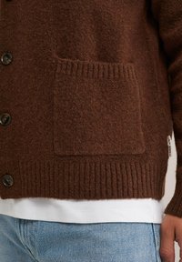 Cardigan marrón texturizado con dobladillo acanalado, cierre de botones y detalle de bolsillo frontal, llevado sobre una camisa blanca y jeans azules.