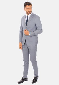 Completo grigio chiaro con vestibilità slim, giacca monopetto, rever a punte e pantaloni coordinati; abbinato a una camicia bianca e una cravatta scura.