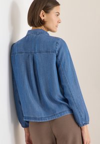 Denim-Oberteil in Mittelblau mit langen Ärmeln, geknöpftem Kragen, elastischen Bündchen und Rückenpasse mit Falten. Weiche Textur und klassisches Design.
