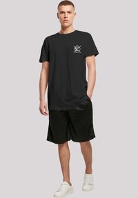 Svart bomull T-skjorte med korte ermer og rund hals, med et hvitt grafisk design på venstre bryst. Kombinert med svarte shorts og hvite joggesko.