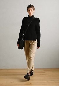 Black ribbed sweater with an asymmetrical neckline, beige tapered trousers, and black shoes. A crossbody bag is worn over the shoulder.