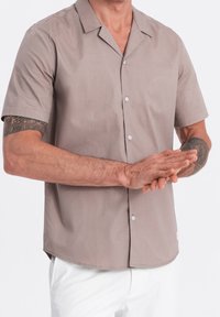 Béžová košile s krátkým rukávem, špičatým límečkem, zapínáním na knoflíky a hladkou texturou. Nahoře jsou nošeny bílé kalhoty s jemným švem.