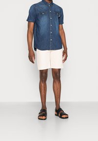 Koszula dżinsowa z krótkim rękawem i dwoma kieszeniami na piersi, zestawiona z jasnobeżowymi szortami i czarnymi sandałami. Swobodny krój z luźnym dopasowaniem.