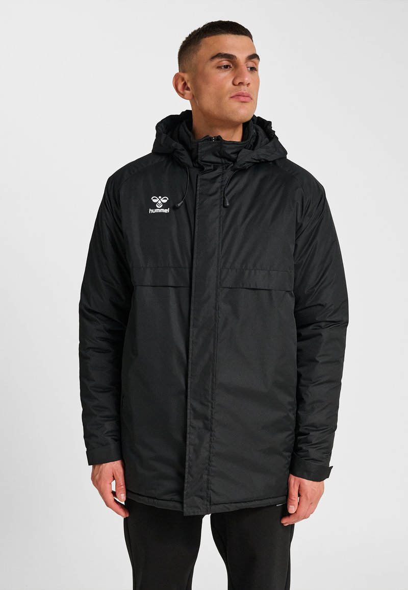 Svart vadderad jacka med huva, med dragkedja framtill, två bröstfickor och en broderad logotyp på vänster sida. Slät textur.