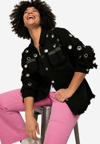 Zwarte denimjas met zilveren oogjes, franjes aan de randen en studs op de zakken, gecombineerd met roze flared broek, tegen een neutrale achtergrond.