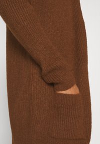 Brun stickad tröja med ribbad textur, som har en sidficka och långa ärmar. Materialet verkar mjukt och varmt.