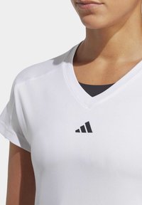 Bílý sportovní tričko s výstřihem do V a krátkými rukávy. Na prsou se nachází černé logo Adidas. Hladká textura, lehký materiál.