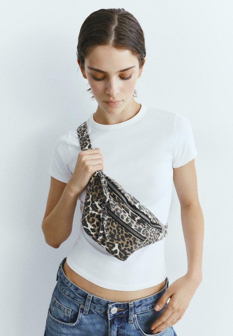 Young woman wearing a white t-shirt and jeans holding a leopard print crossbody bag with two zipper pockets.