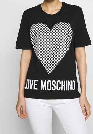 Persoon draagt een zwart T-shirt met een groot wit-zwart geruit hart en de tekst "LOVE MOSCHINO", gecombineerd met een witte broek.