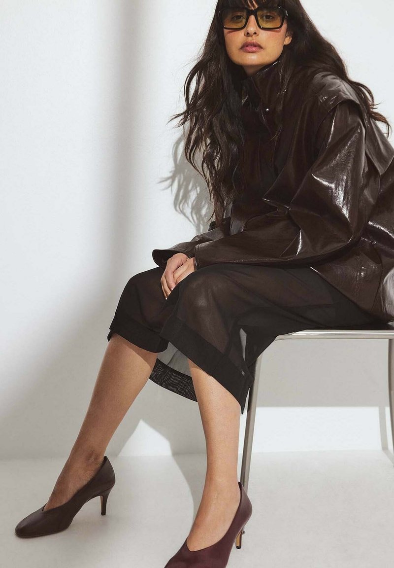 Giacca oversize marrone lucida, gonna nera trasparente con orlo solido e scarpe col tacco bordeaux. Modella seduta su una sedia contro uno sfondo chiaro.