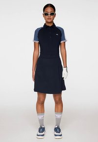 Nainen pukeutuneena tummansiniseen golfasuun, jossa on kauluksellinen paita, hame, valkoinen golfkäsine, urheilulliset aurinkolasit, harmaat sukat ja sinivalkoiset lenkkarit, seisoo suorassa.