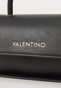 Bolso de mano de cuero negro con una textura suave, que presenta un destacado logo dorado "VALENTINO" en la parte frontal y una forma estructurada.