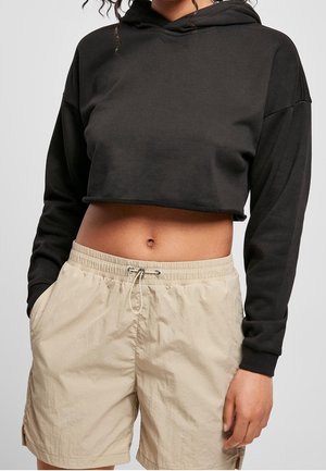 Persoon draagt een zwarte cropped hoodie en beige korte broek met elastische taille en trekkoord, staat met handen in de zakken tegen een effen achtergrond.