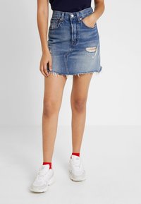 Blue denim mini skirt with frayed hem and distressed details, paired with white sneakers and red ankle socks. Features front pockets and button closure.