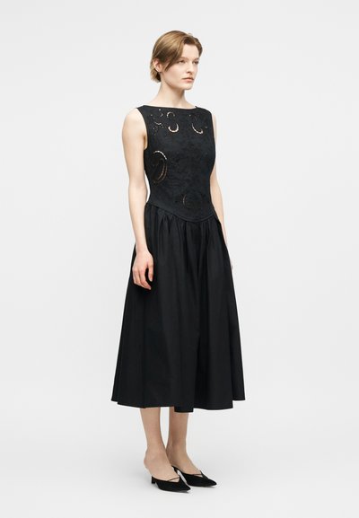 Kobieta stojąca w czarnej sukience bez rękawów z wycięciami na dopasowanym gorsecie i rozkloszowanej spódnicy midi.