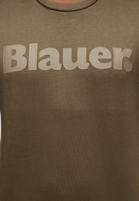 Καφέ μπλούζα με στρογγυλή λαιμόκοψη και μεγάλο μπεζ κείμενο "Blauer." τυπωμένο στο στήθος.