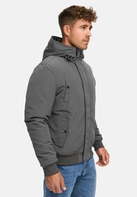 Chaqueta gris acolchada con cierre de cremallera, capucha ajustable y dos bolsillos laterales. Presenta puños acanalados y una textura mate suave.