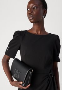 Borsa in pelle nera con logo "DKNY" in rilievo, forma rettangolare elegante, cuciture bianche e tracolla in pelle. Modella che indossa un vestito nero.