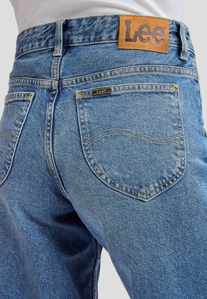 Modré riflové nohavice s zadnými vreckami, ktoré majú žlté prešitie, a hnedou koženou záplatou značky Lee na páse.