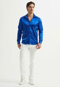 Chemise en satin bleu à manches longues avec un col classique, fermeture boutonnée sur le devant et design ajusté, associée à un pantalon blanc slim-fit et des baskets blanches.