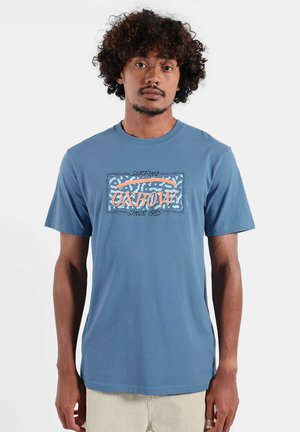 Homme aux cheveux bouclés portant un T-shirt bleu à manches courtes avec le logo "OXBOW Surfing Since 1985" et un pantalon beige, debout devant un fond blanc.