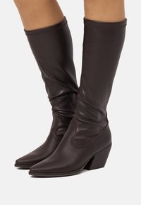 Botas altas hasta la rodilla de cuero marrón con diseño arrugado, punta puntiaguda y tacón apilado. Cuentan con una cremallera lateral para facilitar su uso.