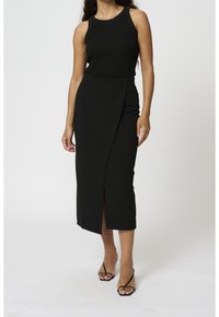 Débardeur noir à côtes associé à une jupe midi noire avec une fente avant et un design asymétrique. Les chaussures sont des sandales à lanières fines.
