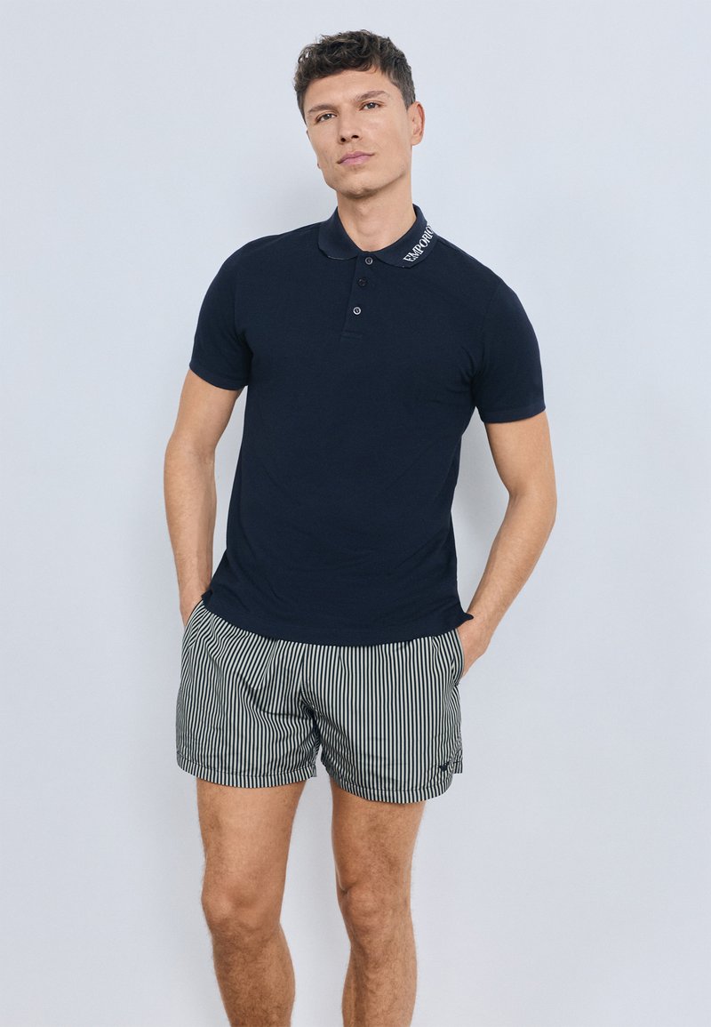 Tamsiai mėlynas polo marškinėliai su dviejų mygtukų siūle ir išsiuvinėtu apykakliu, derinami su dryžuotais šortais. Lygus tekstūra ir prigludęs dizainas.