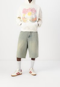 Felpa crema con grafica colorata, pantaloni shorts in denim azzurro chiaro, calzini bianchi e sneakers crema con righe rosse e suole beige.